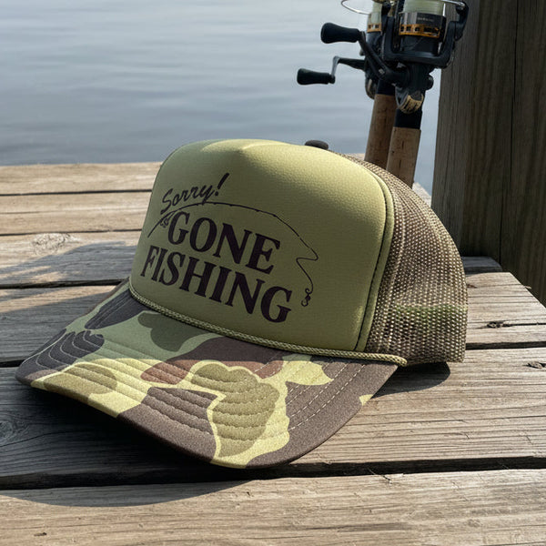 Camouflage fishing cap with 'Sorry! Gone Fishing' text sitting on a wooden dock next to fishing poles