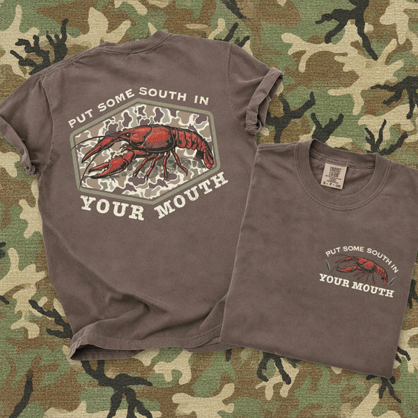 Brown Comfort Colors southern crawfish graphic tee with vintage camo design and “Put Some South In Your Mouth” slogan shown front and back on camo background