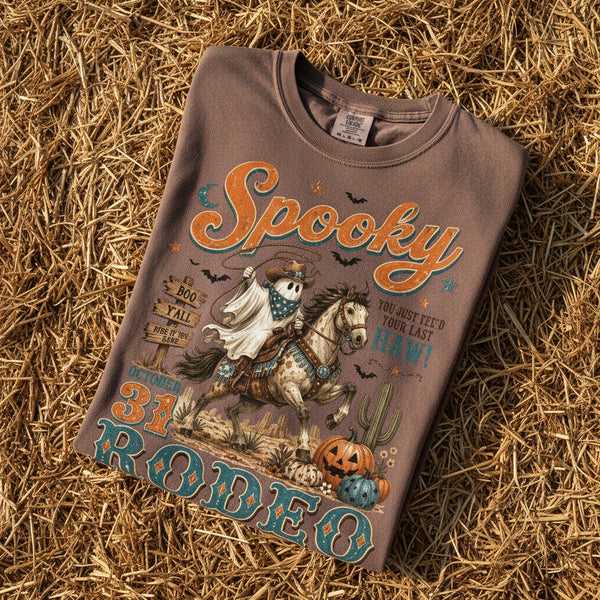 A brown short-sleeve t-shirt with a vintage-style Halloween graphic on the front. The graphic features a ghostly cowboy riding a horse, with the text 'Spooky 31st Robeoo' in orange and teal lettering, along with pumpkins, cacti, and bats.