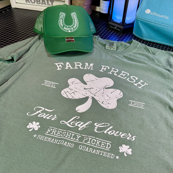 Green t-shirt with 'Farm Fresh Four Leaf Clovers' design and green lucky horseshoe western cap on a table.