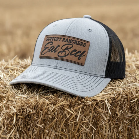 Gray and black cap with "Support Ranchers Eat Beef" leather patch on hay bale