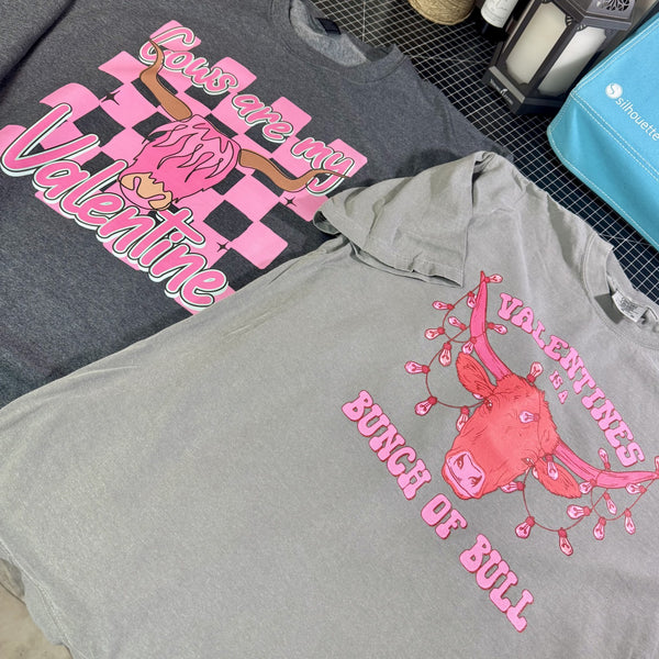 Two western Valentine graphic tees laid flat, featuring pink bull designs and Valentine sayings on gray shirts