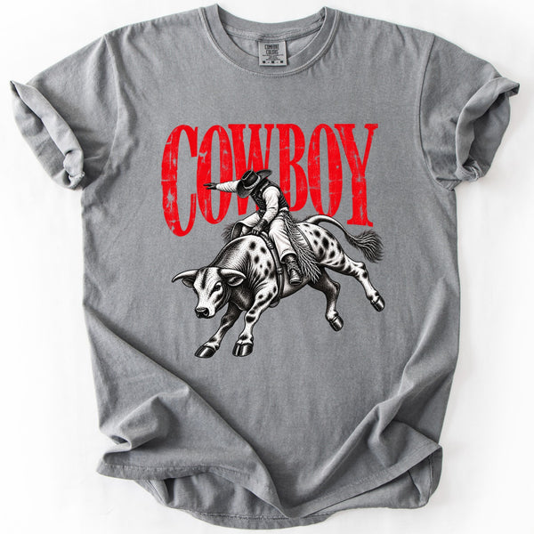 Vintage cowboy bull rider graphic tee on gray Comfort Colors shirt with distressed red COWBOY lettering and black-and-white bucking bull artwork.