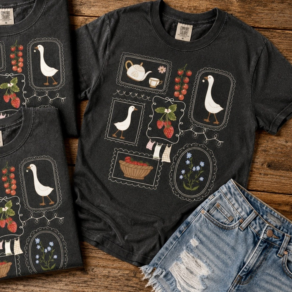 A dark charcoal gray graphic t-shirt featuring a vintage-style tea-time and goose motif, styled in a flat-lay with matching folded t-shirts and distressed denim shorts on a rustic wooden background.