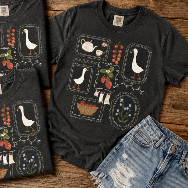 A dark charcoal gray graphic t-shirt featuring a vintage-style tea-time and goose motif, styled in a flat-lay with matching folded t-shirts and distressed denim shorts on a rustic wooden background.