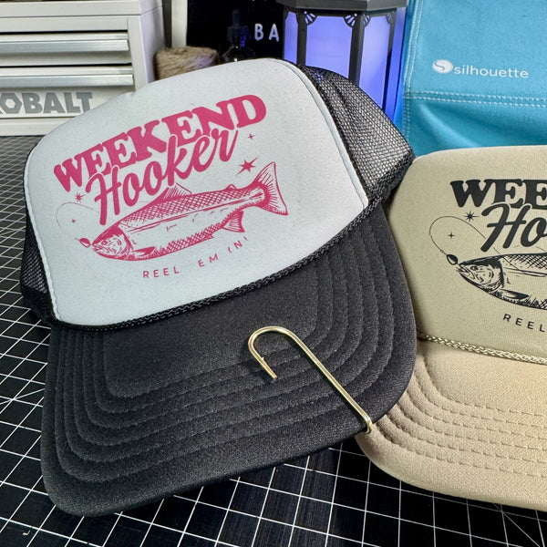 Two trucker hats with 'Weekend Hooker' design on a grid surface.
