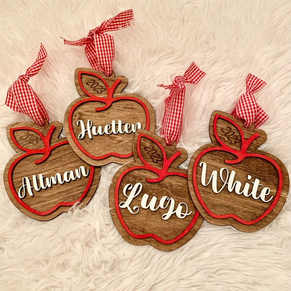 Four personalized wooden apple ornaments with red painted outlines and red-and-white gingham ribbon hangers, arranged on a white faux-fur surface. The ornaments are engraved with names (Allman, Huetter, Lugo, White) and years (2025).