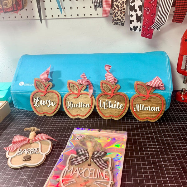 Four personalized wooden apple ornaments with red gingham bows are displayed on a cutting mat in front of a turquoise pencil case. 