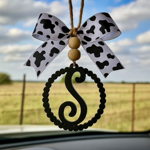 Decorative hanging letter ornament on rear view mirror with a cow print bow and letter 'S' against a cloudy sky.