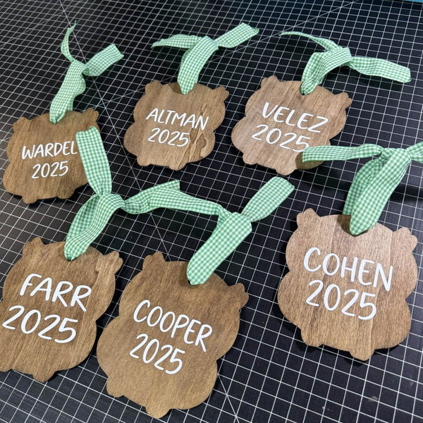 Wooden tags with names and '2025' printed on them, attached to green ribbons, on a grid background.