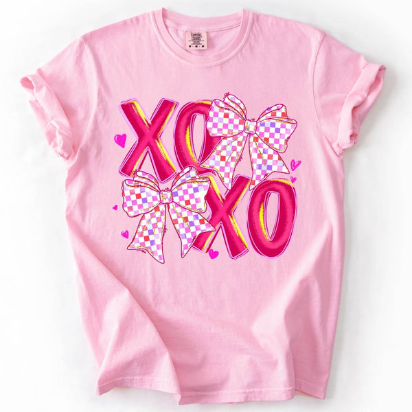 Pink Comfort Colors Valentine’s Day t-shirt featuring bold XOXO lettering with pink checkered bows and heart accents
