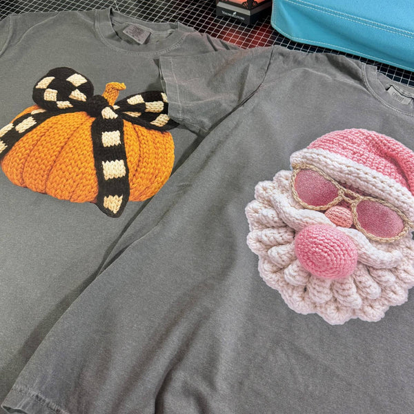 Gray t-shirt with a knitted pumpkin and pink knitted pink Santa holiday graphic design on a checkered background.