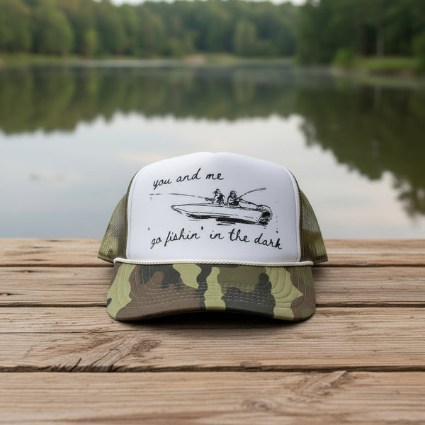 Camo trucker hat with sketch of couple fishing in a boat and text ‘you and me go fishin’ in the dark.
